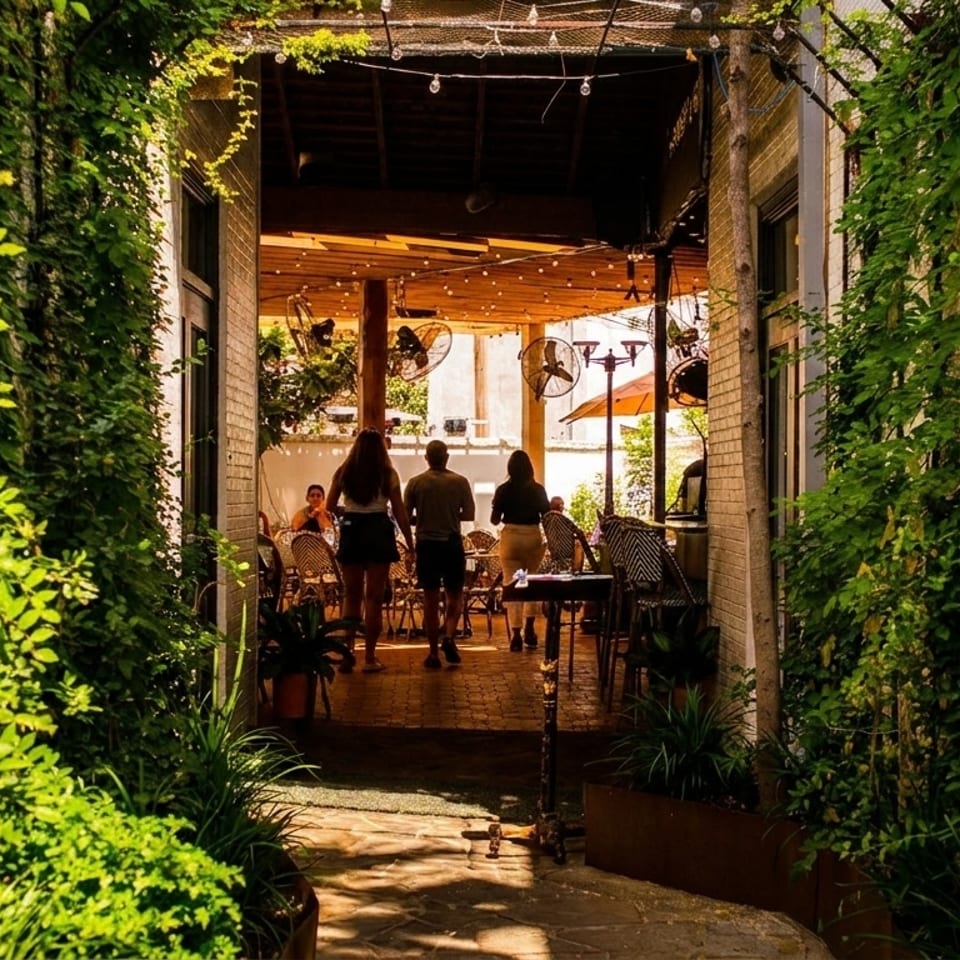 People walking into a restaurant patio area, framed by lush green vines and string lights.