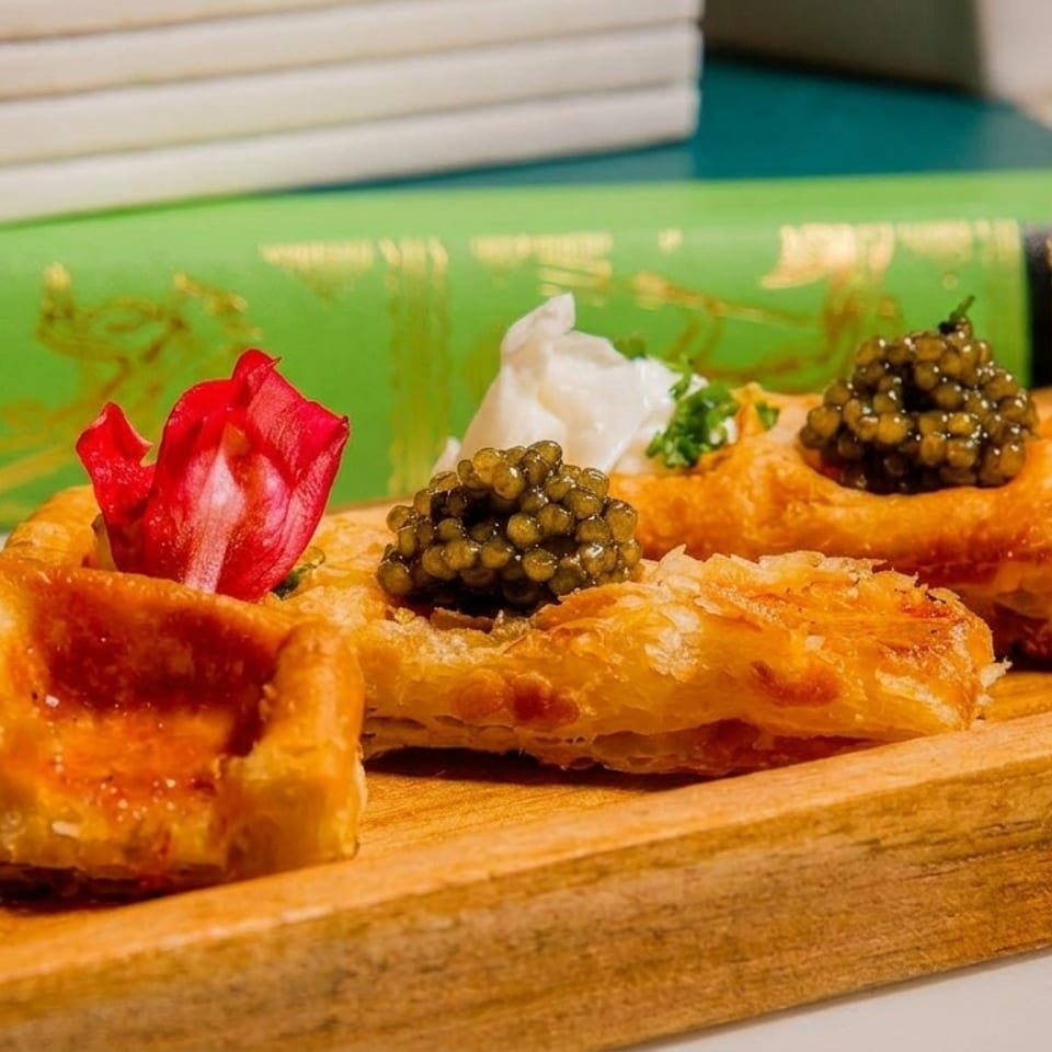 Golden puff pastry appetizers topped with caviar, sour cream, and rose petals on a wooden tray.